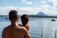 Imagem mostra homem com criança no colo. Ambos olham para o mar.