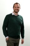 Homem de blusa verde, mão no bolso e sorrindo