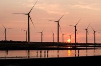Wind turbines silhouetted against a sunset, reflecting in calm water as people walk by.