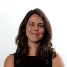 Mulher de cabelos castanhos, de braços cruzados e sorrindo