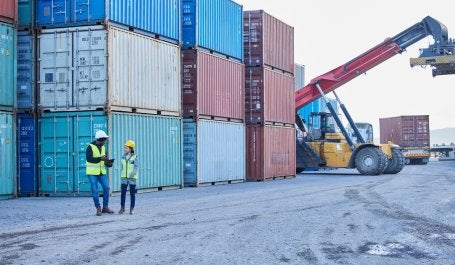 pessoas caminha próximas a containeres em área portuária