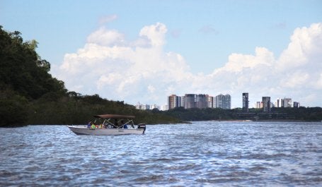 A paisagem mostra um rio, com um barco navegando, mata em volta e ao fundo prédios da cidade de Manaus