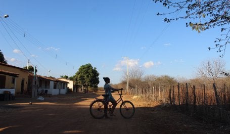 Imagem mostra mulher em uma bicicleta e, ao fundo, paisagem de Caatinga