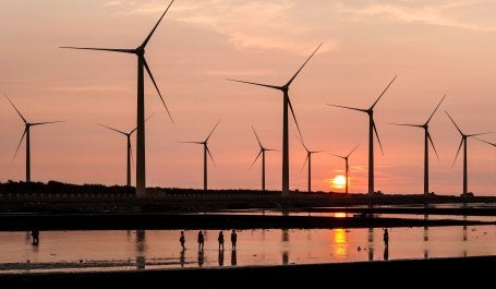 Wind turbines silhouetted against a sunset, reflecting in calm water as people walk by.