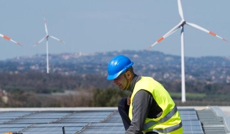 homem durante instalação de painel solar com estações de energia eólica ao fundo