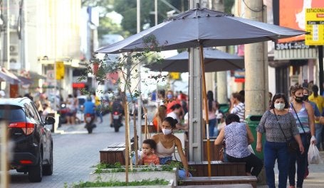 foto de uma rua em São José dos Campos. Pessoas conversam sentadas sob ombrelones na calçada. Carros passam lentamente na rua.